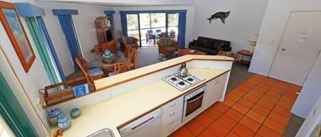 Une cuisine compacte équipée d’une cuisinière à gaz, d’un four et d’un lave-vaisselle, un coin repas avec des chaises et une grande fenêtre agrémentée de rideaux bleus.