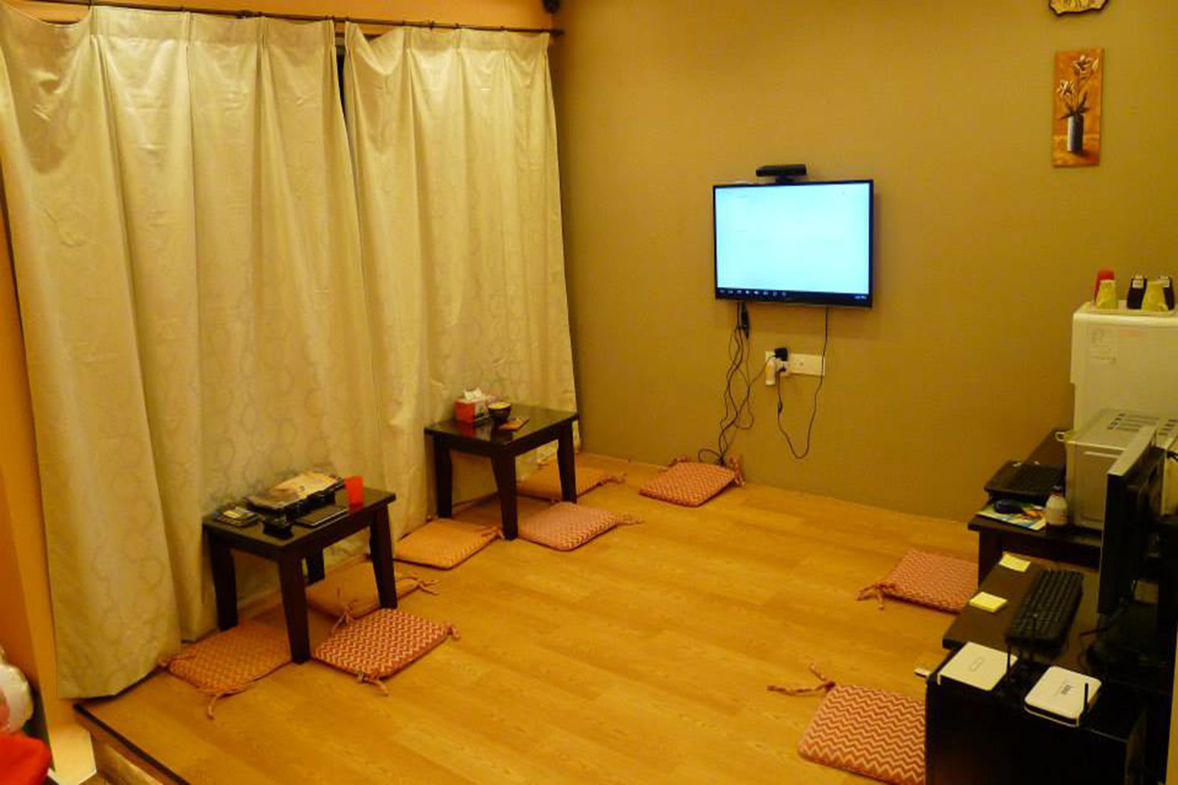 A room with a wooden floor, a television mounted on the wall, a desk with a computer, and a small table with a chair.