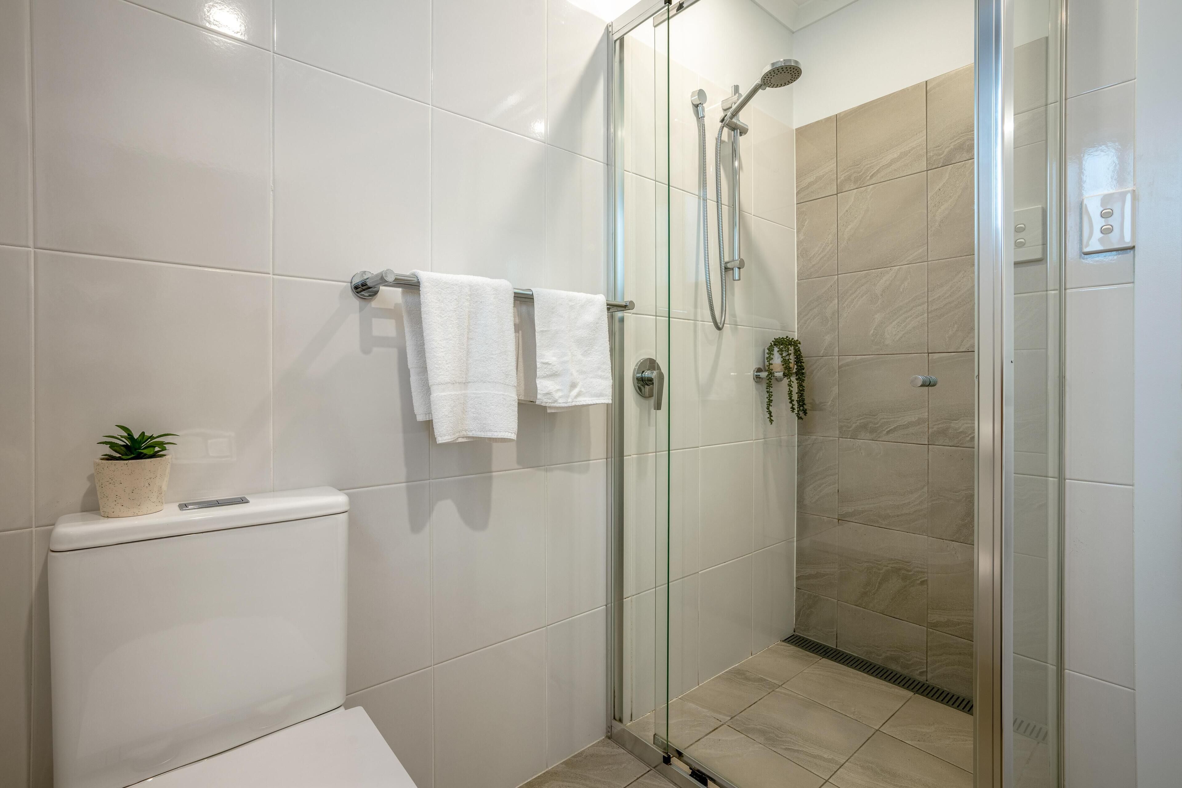 Une salle de bain moderne avec une douche à parois de verre, des carreaux blancs, des toilettes et une petite plante posée sur le réservoir des toilettes.