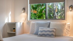 Un dormitorio con una cama grande, una ventana que da a árboles, una mesita de noche con una lámpara y un pequeño estante con una planta.