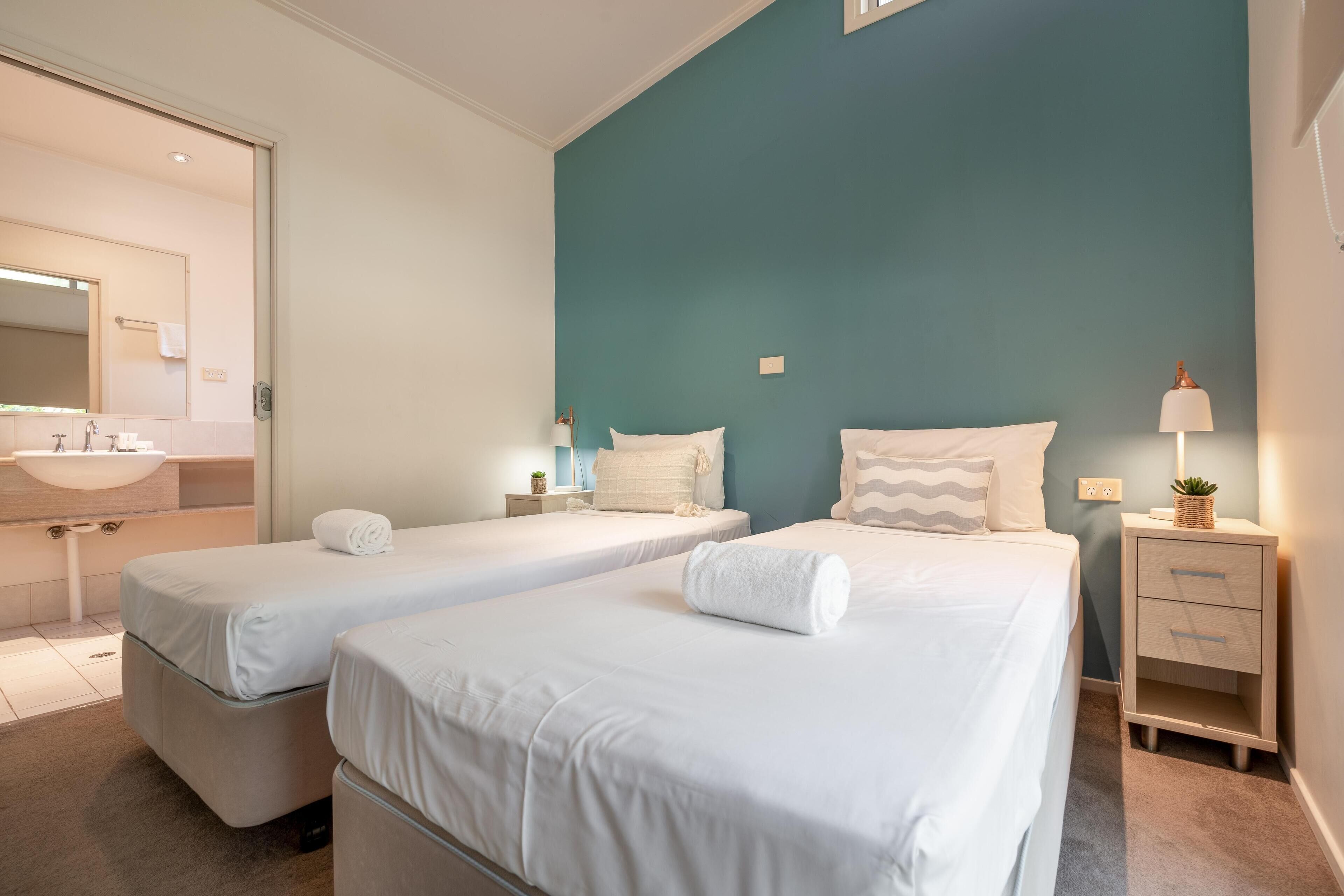 A hotel room with two single beds, a bedside table with a lamp, a nightstand, a mirror, and a bathroom with a sink and towel rack.