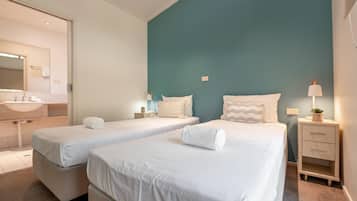A hotel room with two single beds, a bedside table with a lamp, a nightstand, a mirror, and a bathroom with a sink and towel rack.