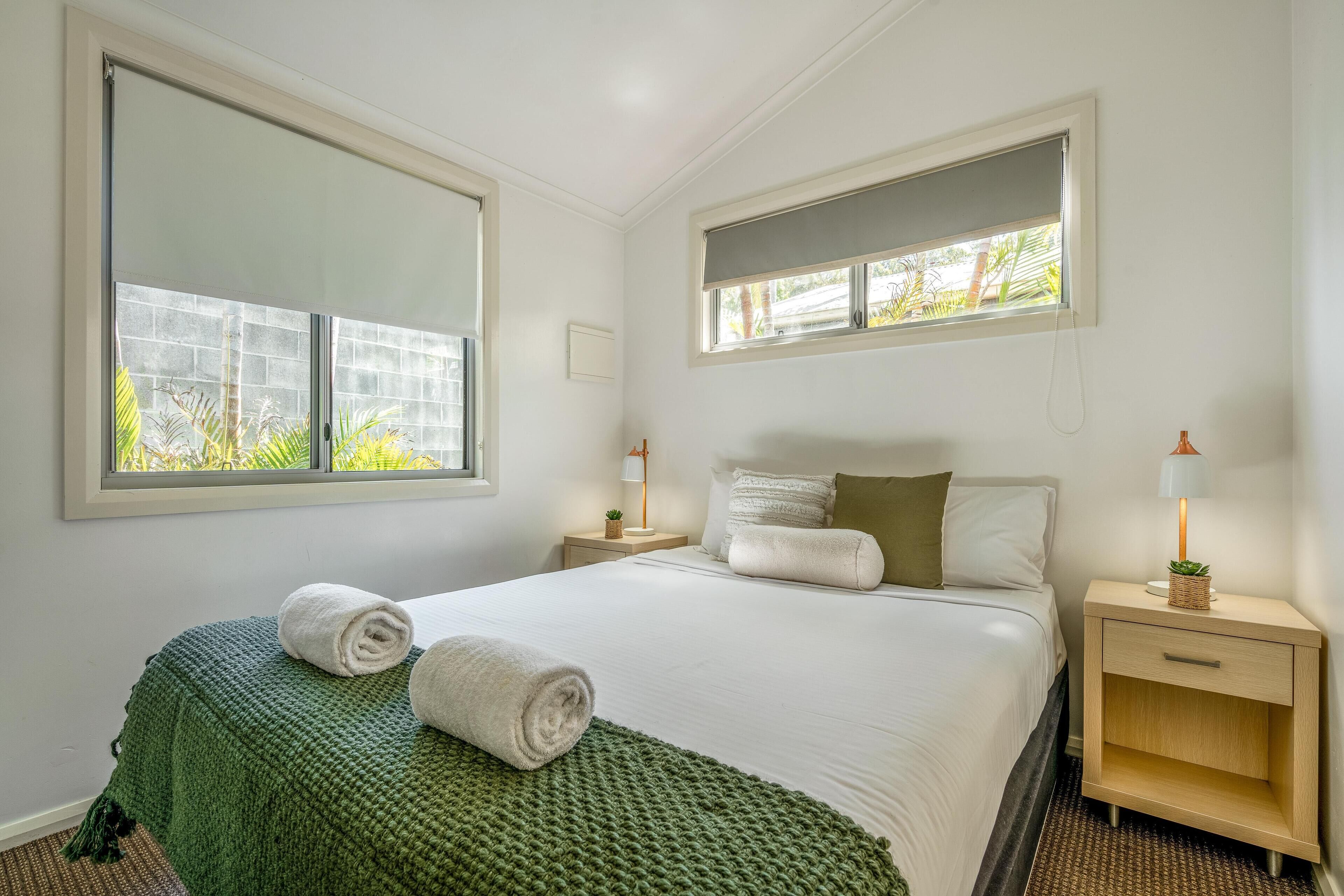 Un dormitorio ordenado con una cama grande, dos mesitas de noche, un ventanal con vistas y un plaid verde de punto sobre la cama.