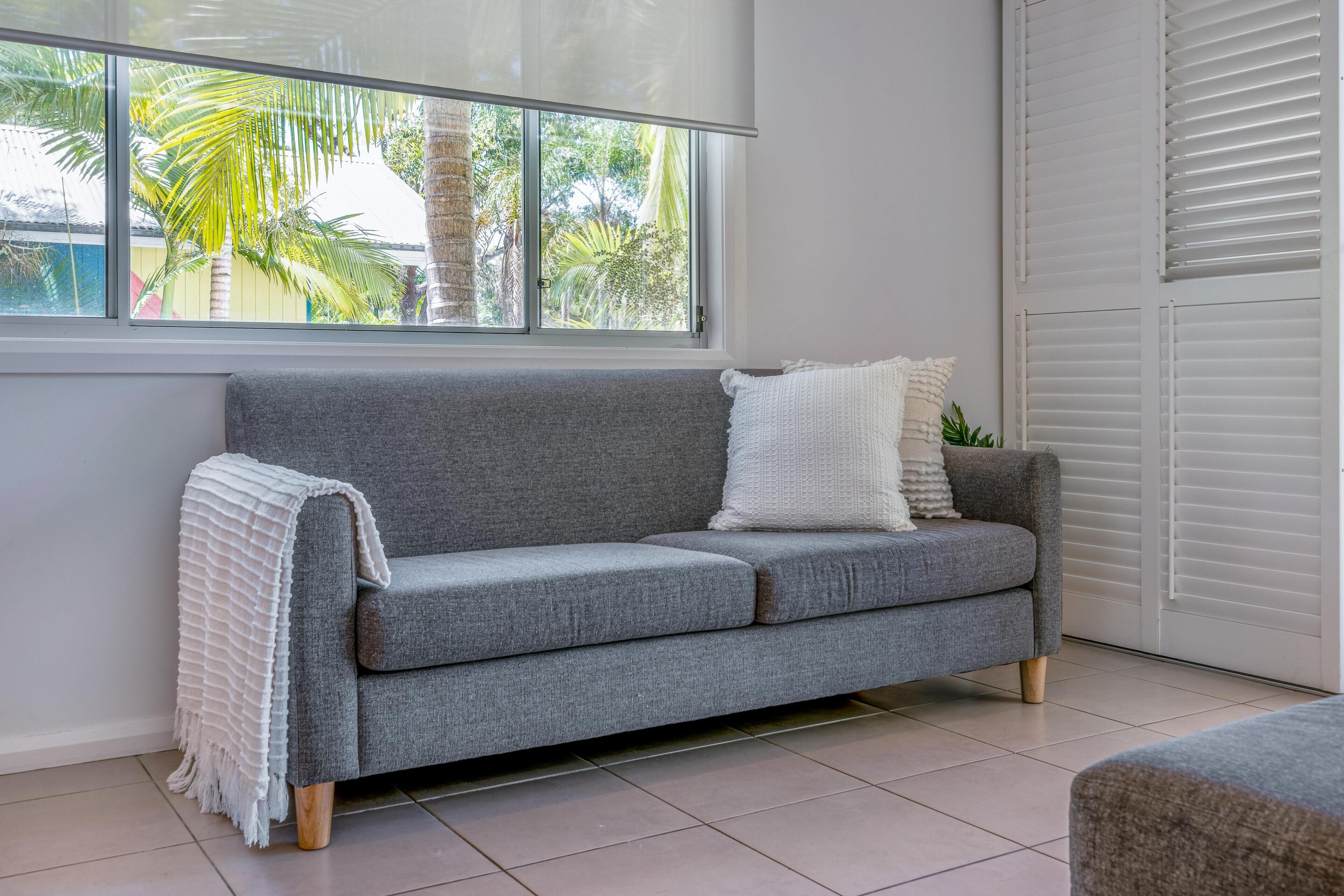 Un sofá gris con una manta blanca y almohadas, situado junto a una ventana con vistas a palmeras.