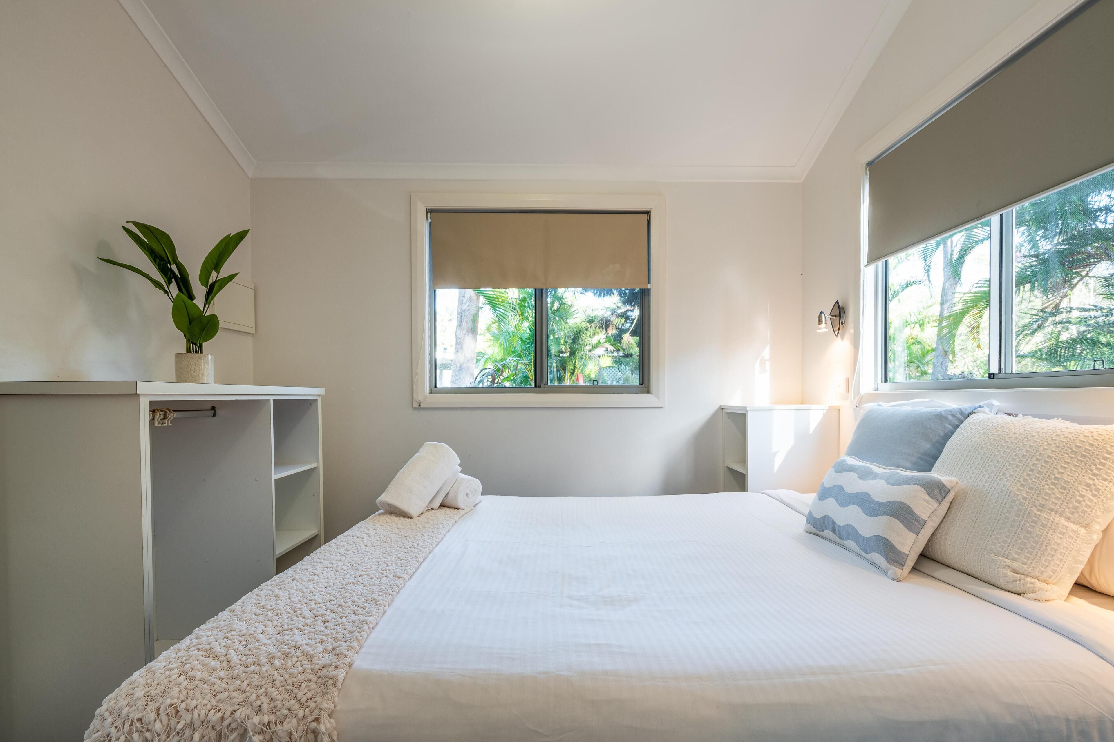 Un dormitorio con cama, ventana, planta y mesita de noche.