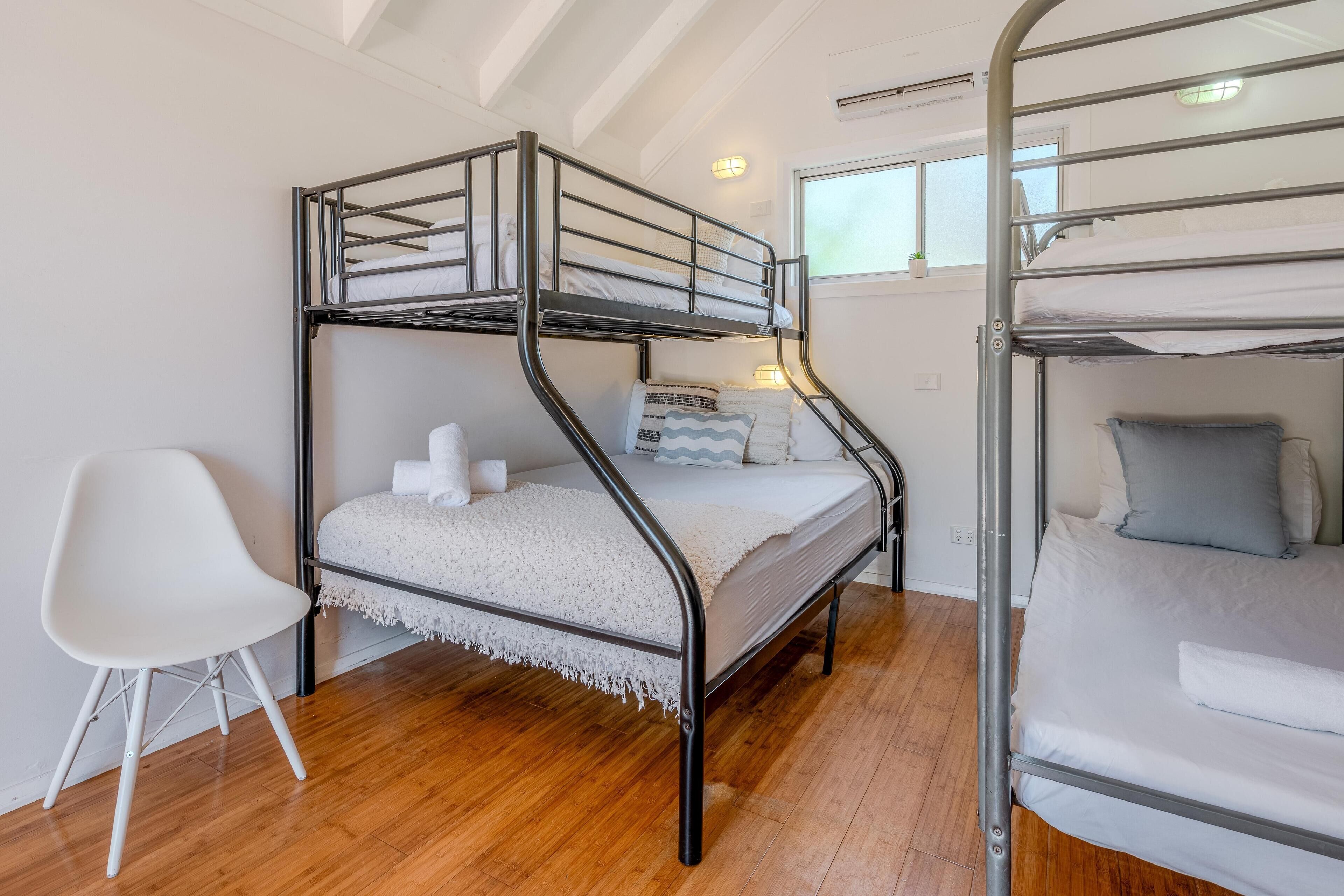 A bunk bed room with a white chair, wooden flooring, and a window providing natural light.