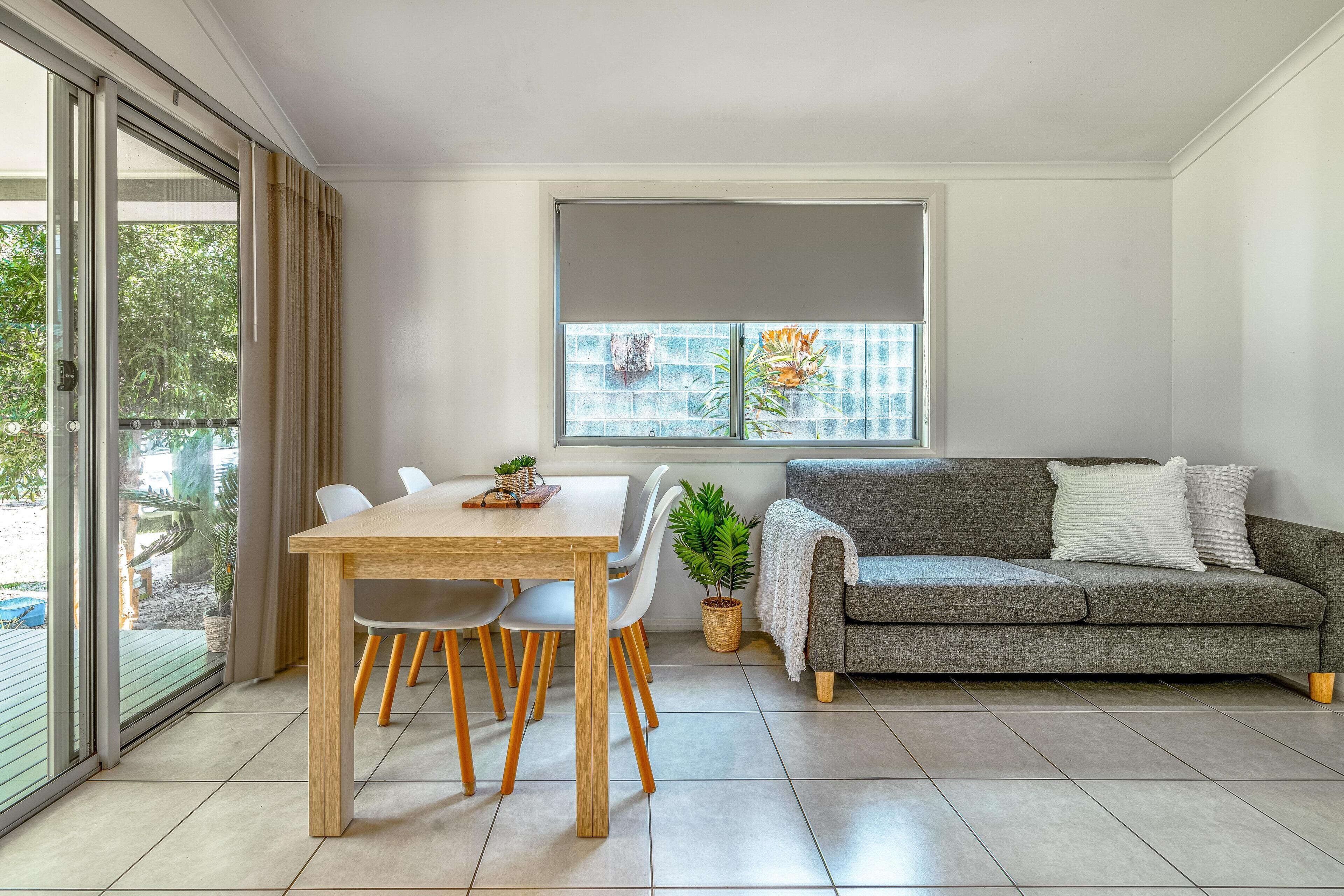 Un salon moderne avec un canapé gris, une table à manger en bois et des chaises, une grande fenêtre donnant sur un espace verdoyant, et une porte-fenêtre coulissante qui mène à un espace extérieur.