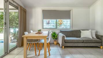 Un salon moderne avec un canapé gris, une table à manger en bois et des chaises, une grande fenêtre donnant sur un espace verdoyant, et une porte-fenêtre coulissante qui mène à un espace extérieur.