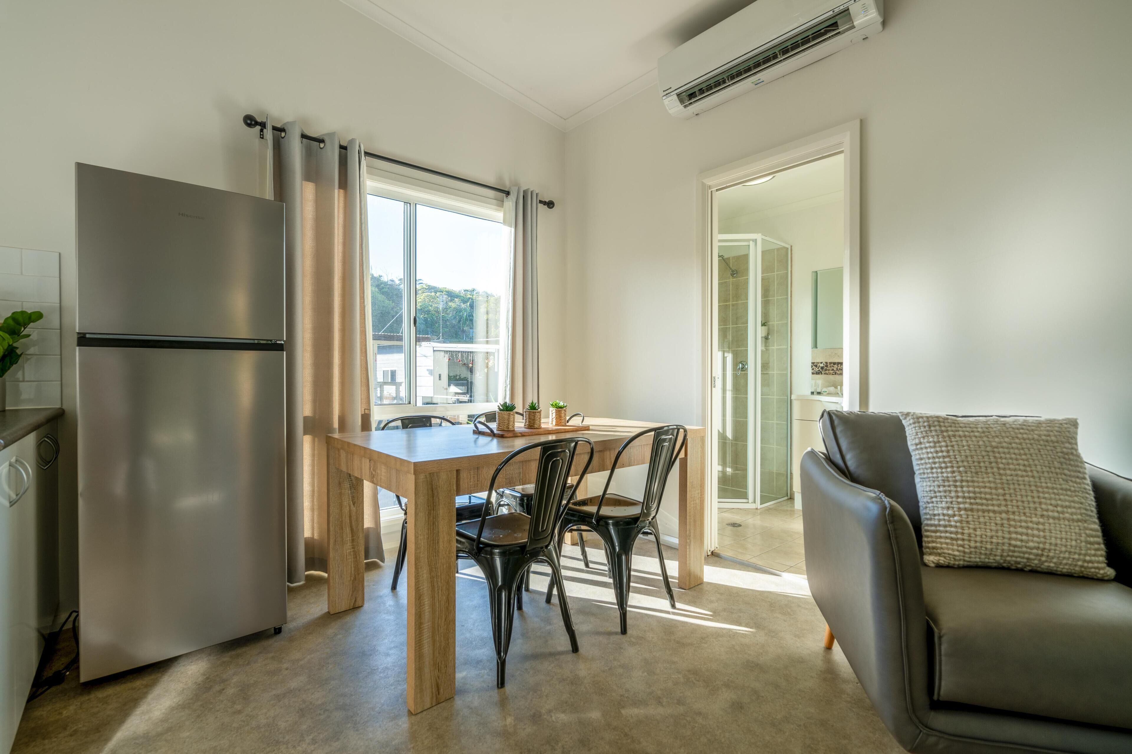 Une cuisine compacte équipée d’un réfrigérateur, d’une table à manger en bois et de chaises. Un canapé est placé près de la fenêtre, et un climatiseur est fixé au mur.