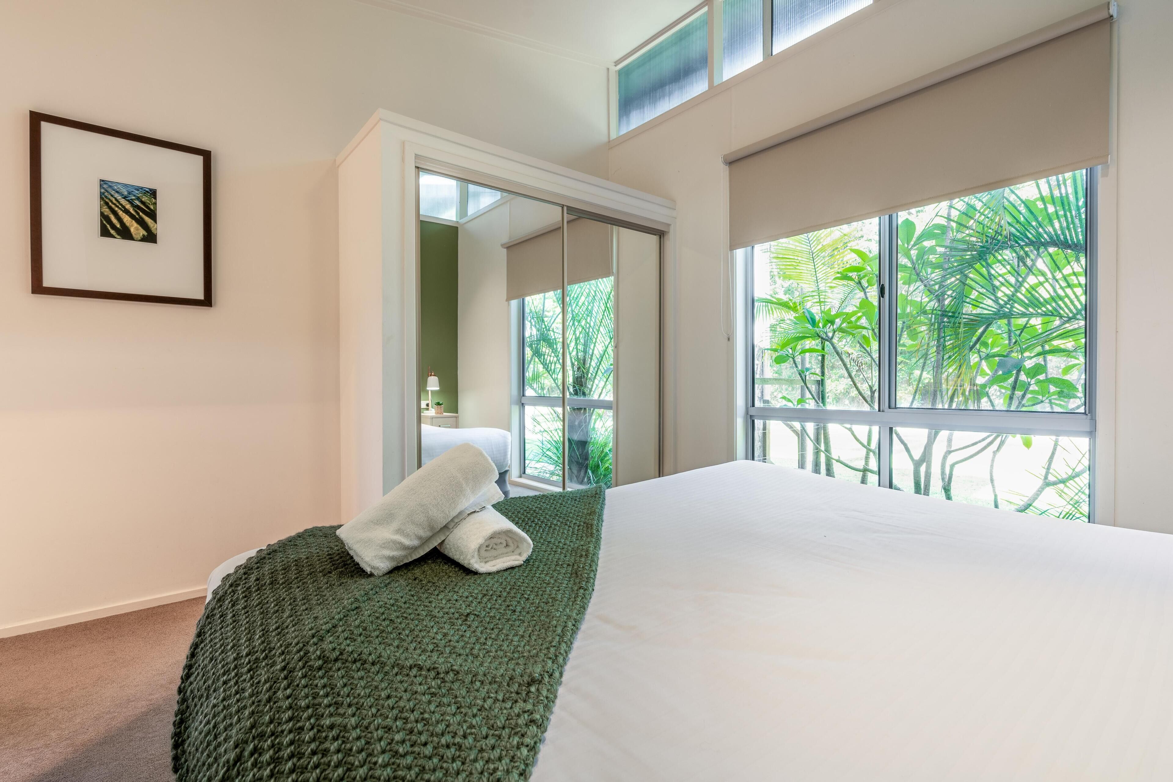 Habitación de hotel con una cama grande, una manta verde, un cuadro en la pared y vistas a la vegetación por la ventana.
