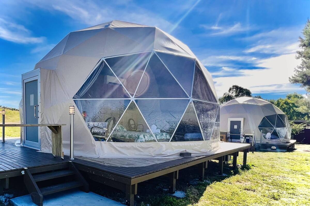 A geodesic dome structure with a wooden deck and a door, set in a grassy area under a clear sky.