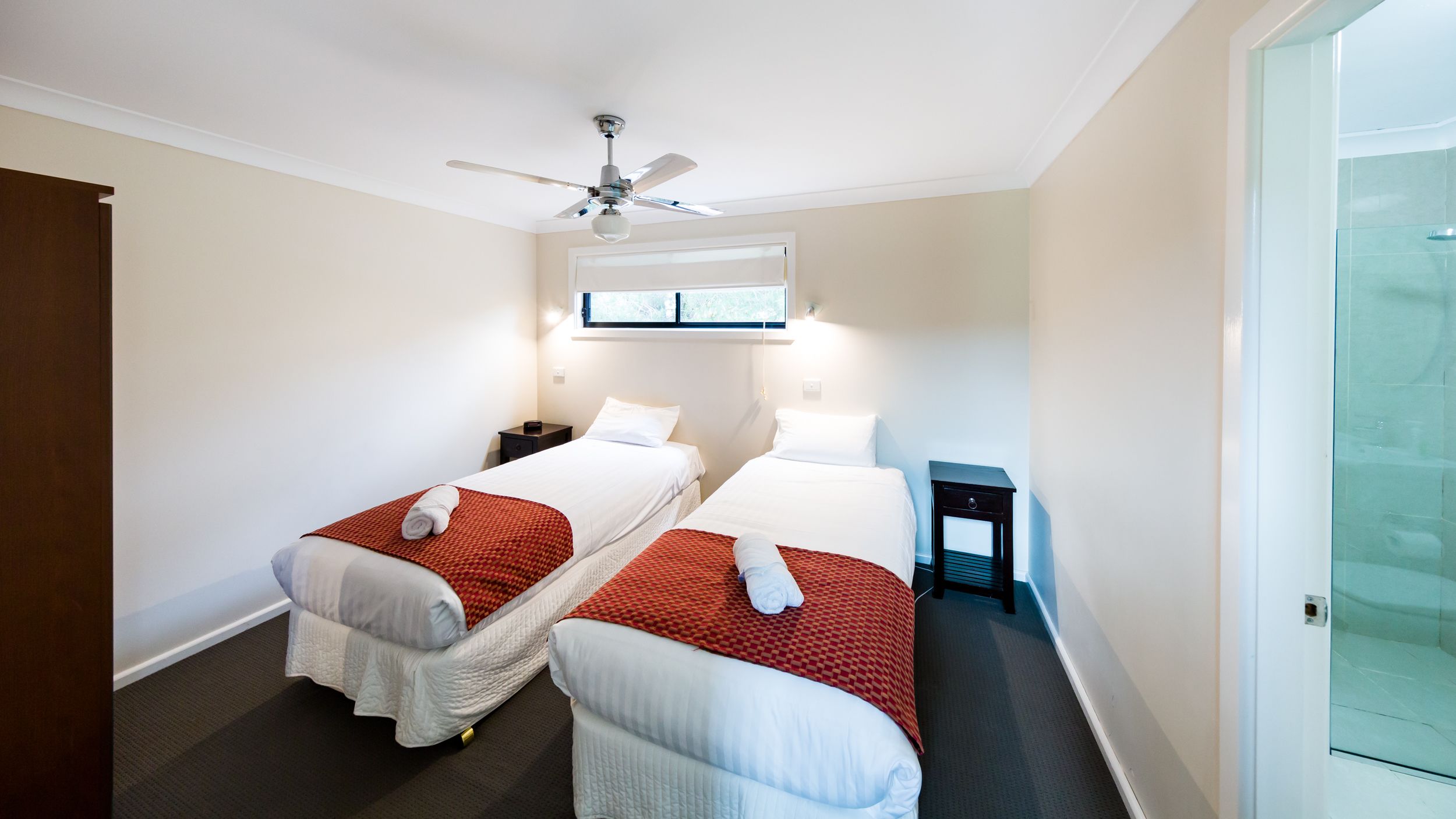 A hotel room with two beds, a ceiling fan, a window with blinds, and a glass shower enclosure.
