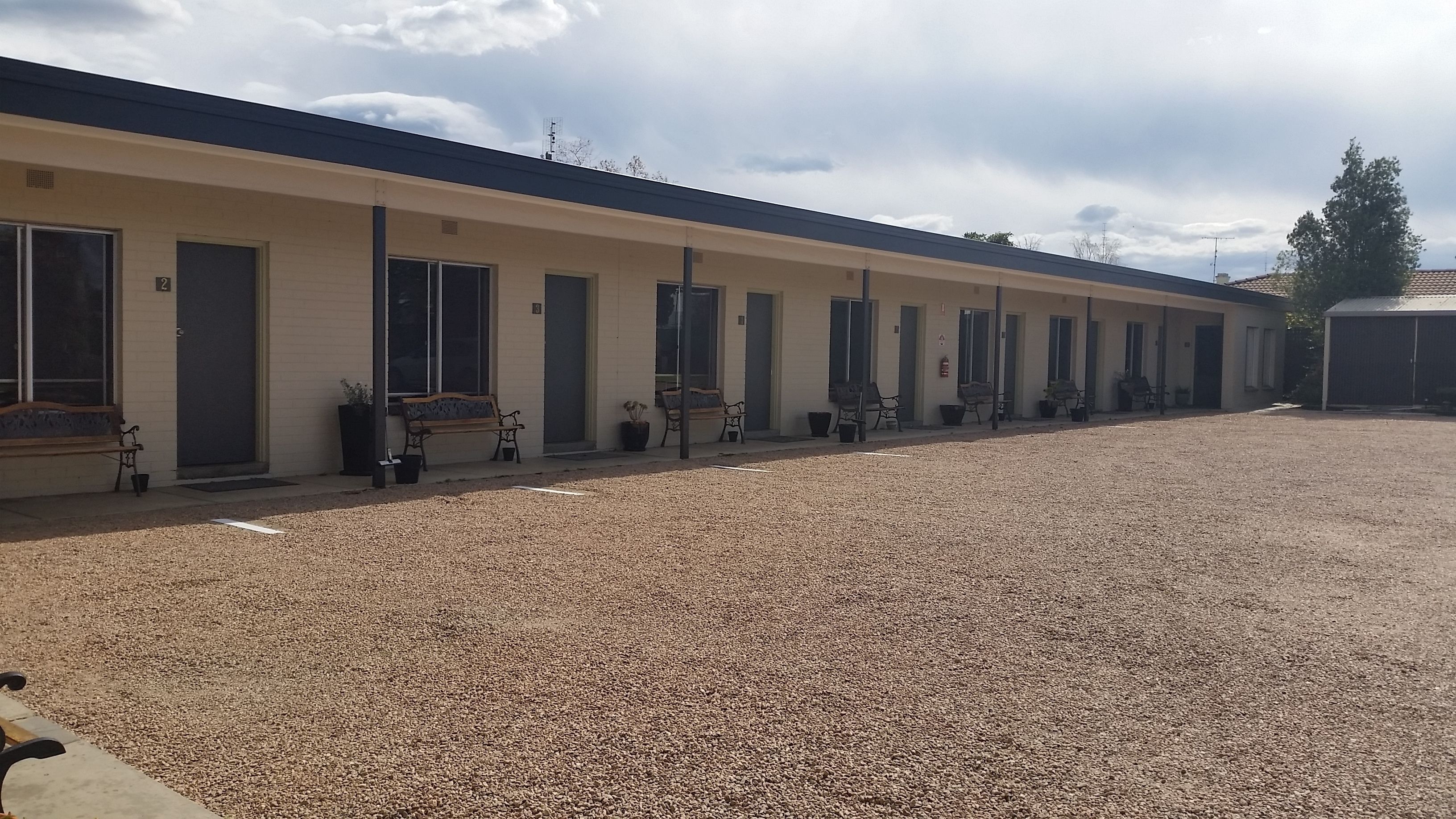 A motel with multiple rooms, each with a door and windows. There are benches and potted plants outside the rooms.