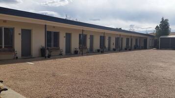A motel with multiple rooms, each with a door and windows. There are benches and potted plants outside the rooms.
