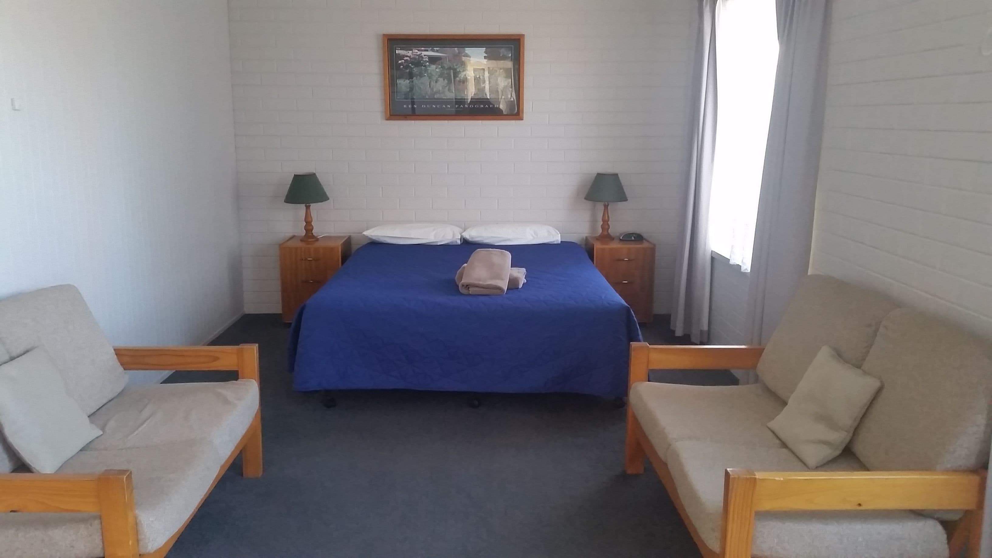 A hotel room with a bed, two nightstands with lamps, a framed picture on the wall, and two armchairs.