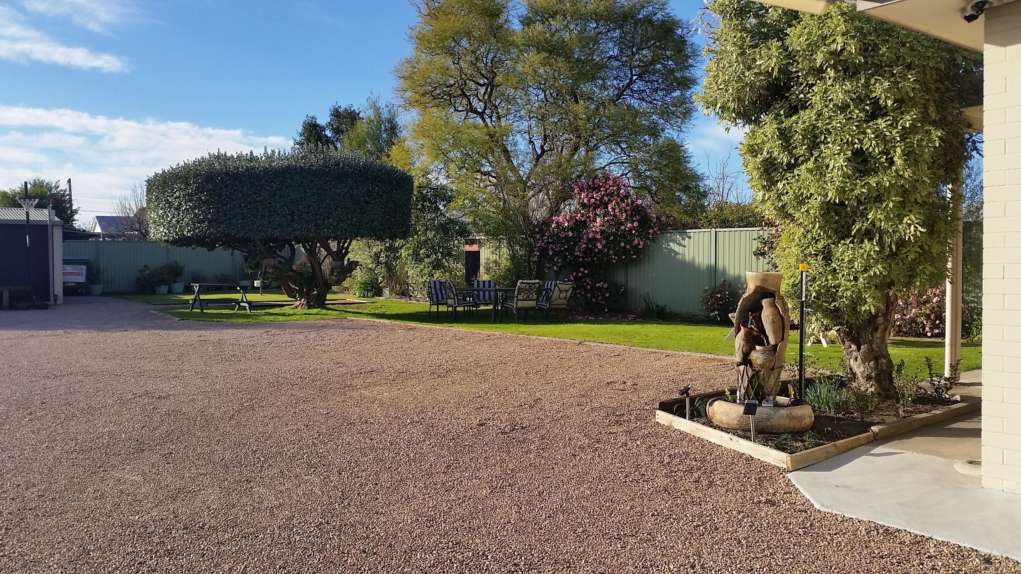 A gravel driveway leading to a house with a garden, a patio with chairs, and a large tree with pink flowers.