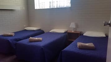 Habitación con tres camas individuales, cada una con almohada y una toalla enrollada. Hay una mesita de noche con lámpara entre las camas.