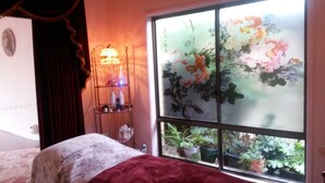 Un dormitorio con cama, una mesita de noche con lámpara, una ventana con vistas a un jardín floral y una cortina.
