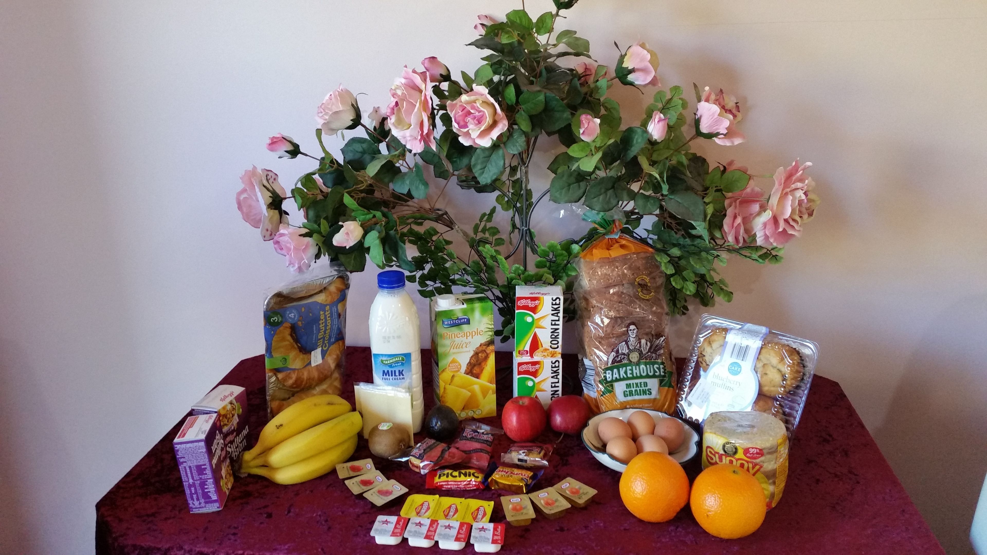 A table with various food items, including bread, milk, apples, oranges, and a plant with pink flowers.