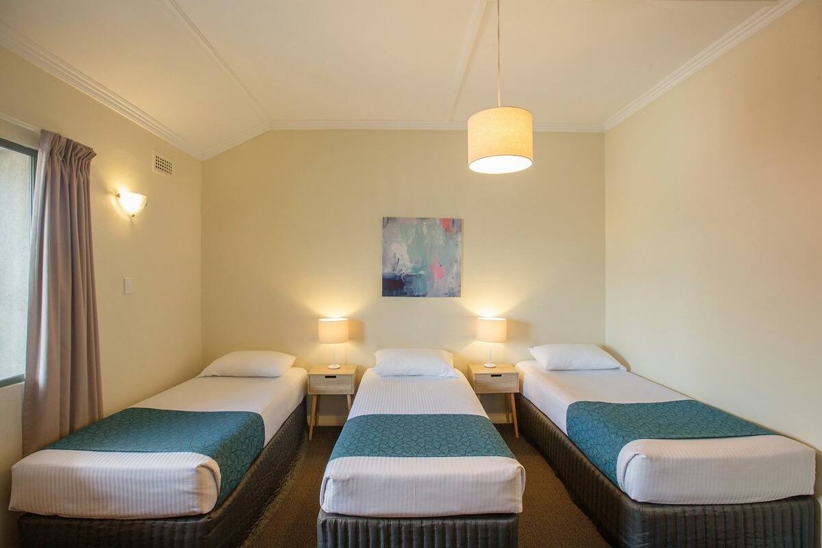 Habitación de hotel con tres camas individuales, cada una con mesita de noche y lámpara, un ventanal grande con cortinas y un cuadro en la pared.
