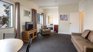 Habitación de hotel con televisión, sofá, silla, mesa de comedor y cama al fondo.