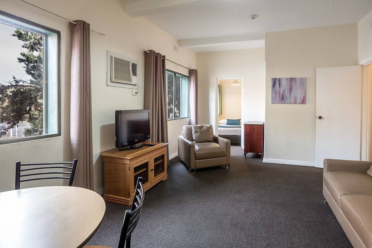 Habitación de hotel con televisión, sofá, silla, mesa de comedor y cama al fondo.