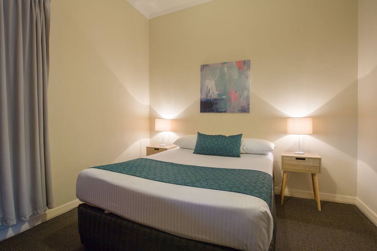 Una habitación de hotel con una cama, dos lámparas de noche, un cuadro en la pared y una ventana con cortinas.