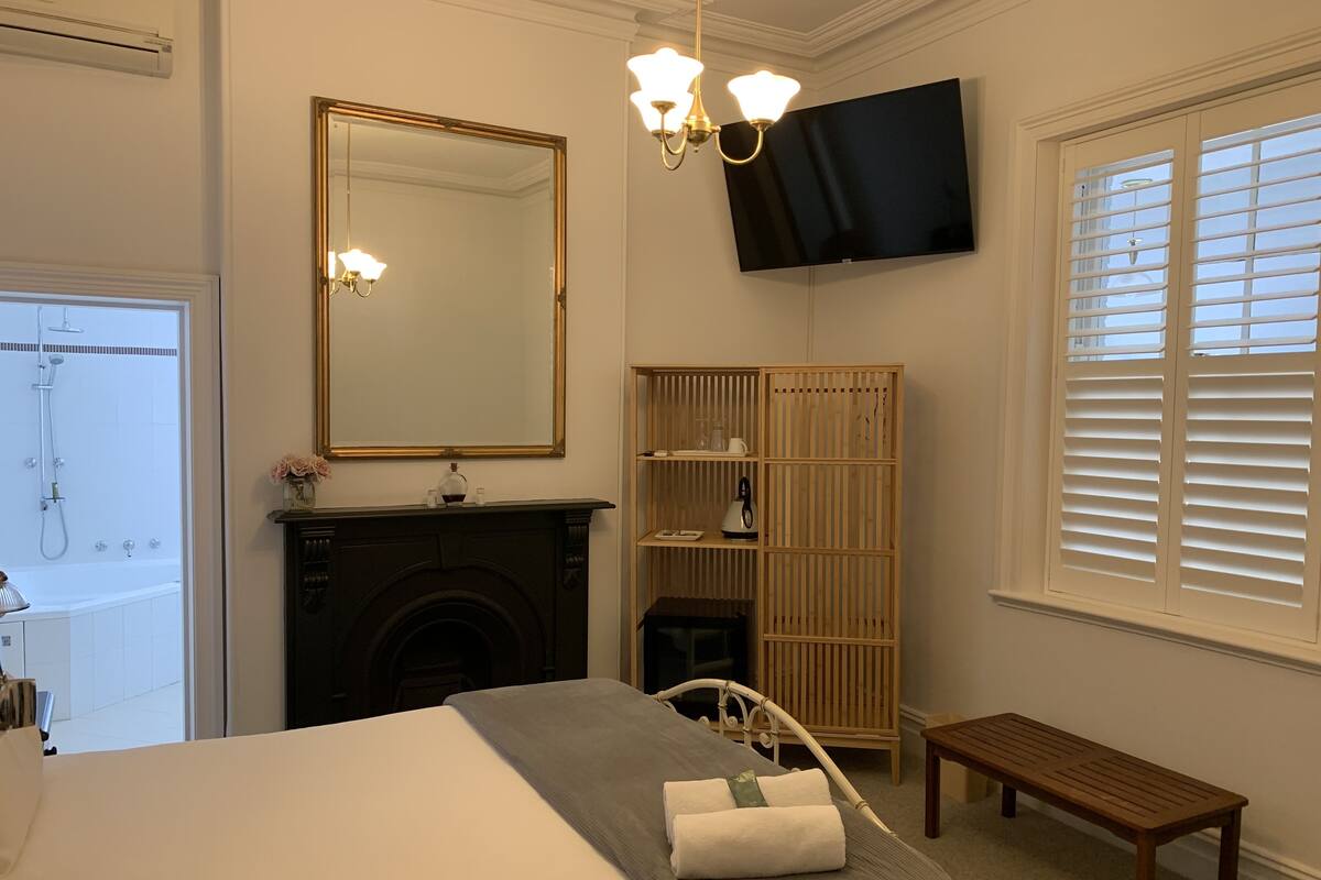 A bedroom with a bed, a television mounted on the wall, a fireplace, a wooden cabinet, and a window with blinds.