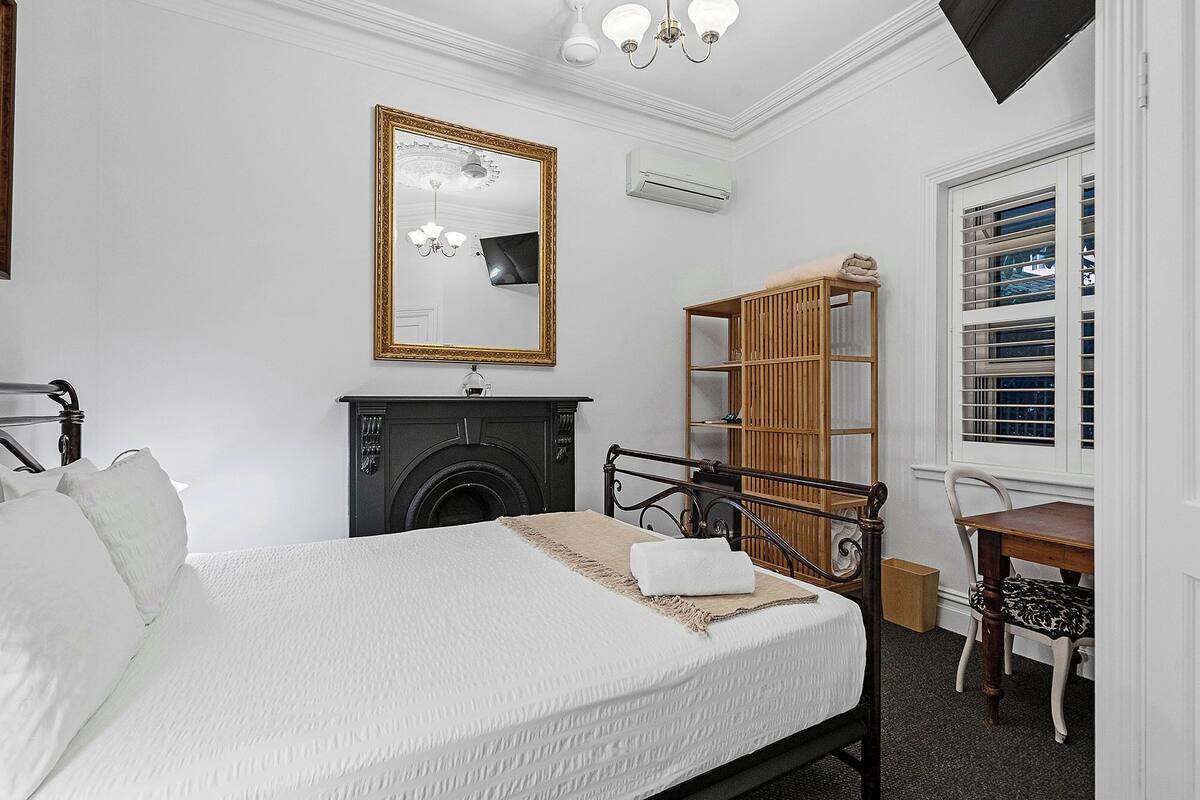 A bedroom with a bed, a mirror, a fireplace, a wardrobe, and a window with blinds.