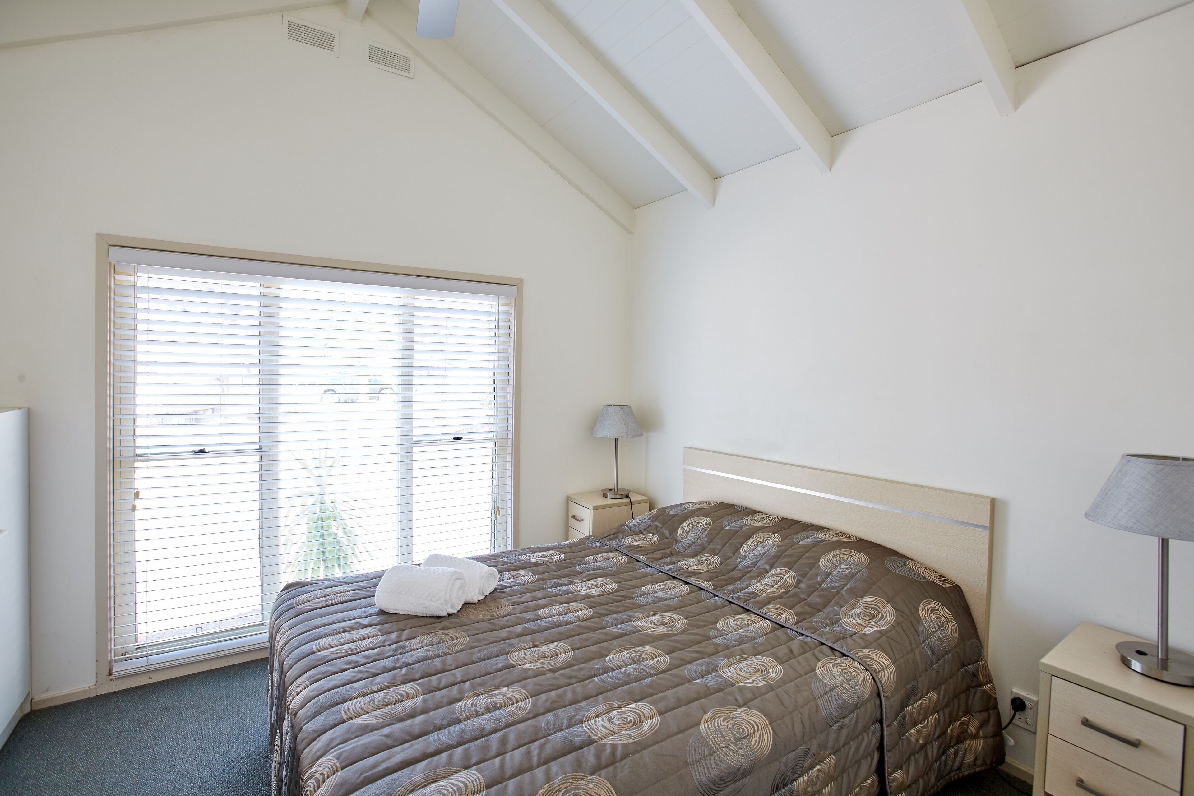 A bedroom with a bed, a window with blinds, a bedside table with a lamp, and a nightstand.