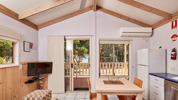 Ein gemütliches Zimmer mit Holzbalkendecke, einem Fernseher auf einem Holzbodenregal, einem Esstisch mit Stühlen, einer Küchenzeile inklusive Kühlschrank und einer Glastür, die auf eine Außenterrasse führt.