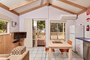 Une pièce chaleureuse avec un plafond en bois, un téléviseur posé sur une étagère en bois, une table à manger avec des chaises, un coin cuisine avec un réfrigérateur et une porte-fenêtre coulissante donnant sur une terrasse extérieure.