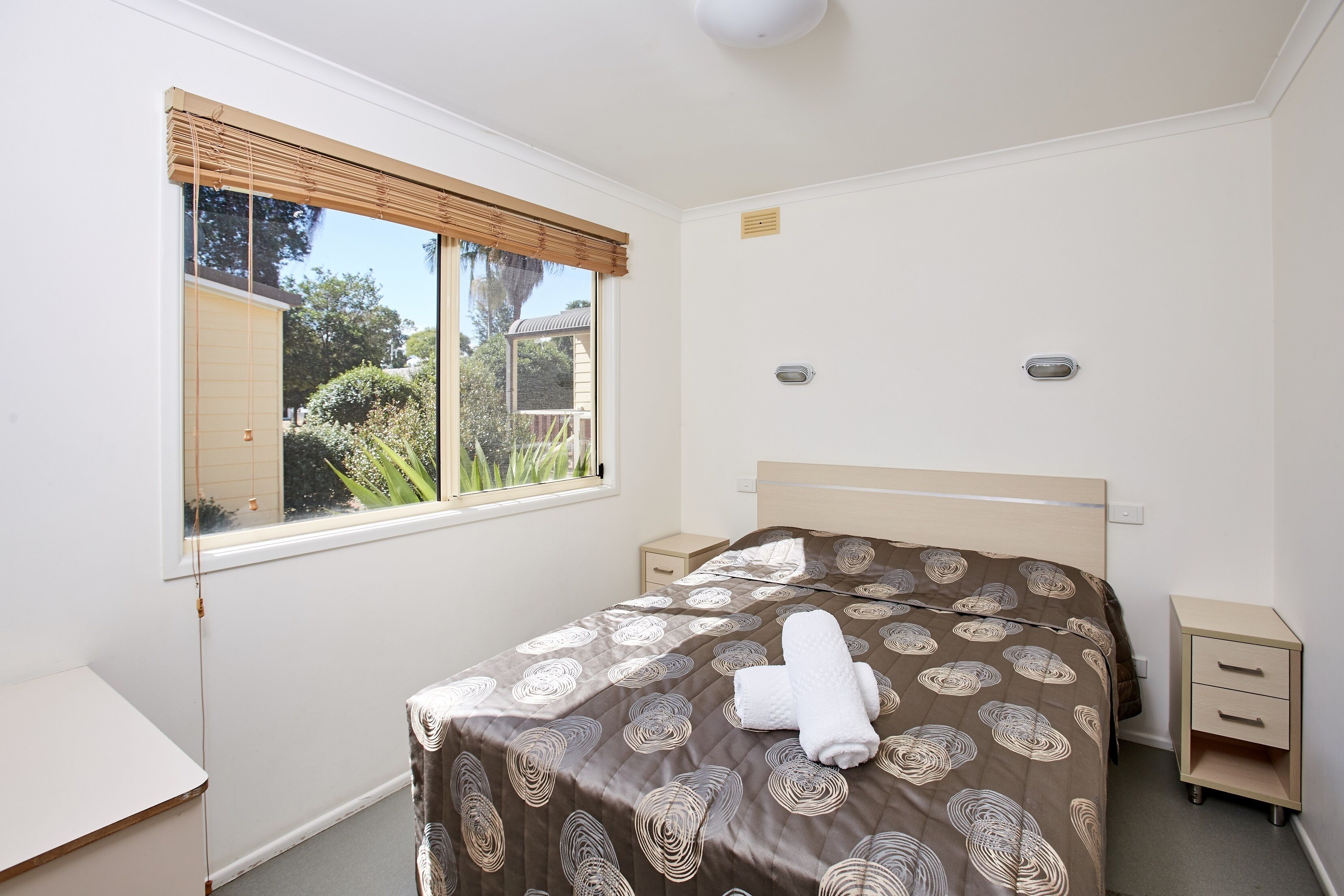 A bedroom with a bed, a bedside table, a nightstand, a window with blinds, and a view of greenery outside.