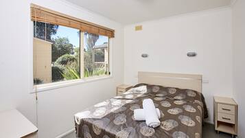 A bedroom with a bed, a bedside table, a nightstand, a window with blinds, and a view of greenery outside.