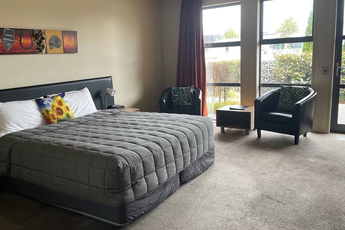 A hotel room with a large bed, a chair, a nightstand, and a window with a view of greenery.