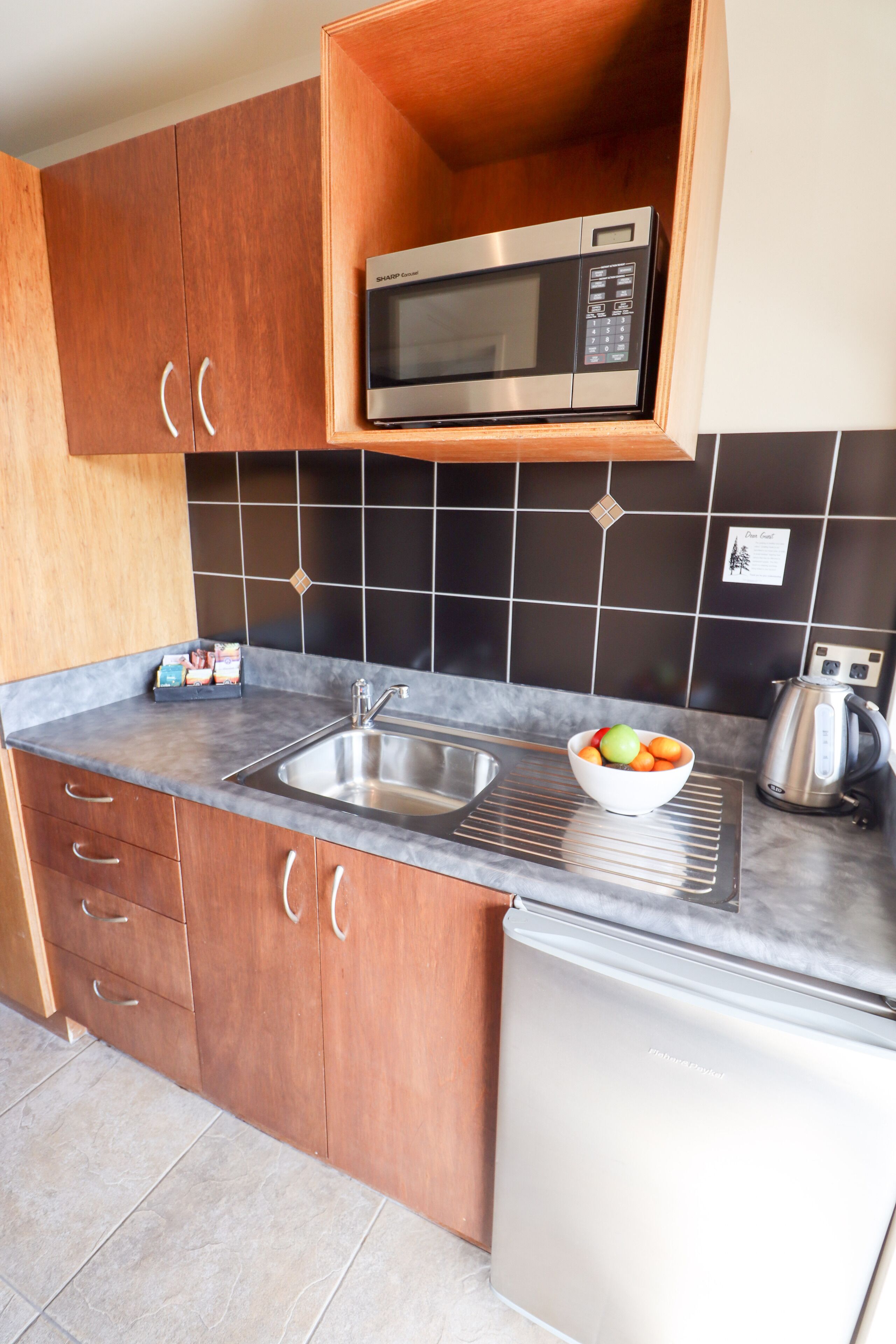 A compact kitchen with wooden cabinets, a stainless steel microwave, a sink, and a kettle.