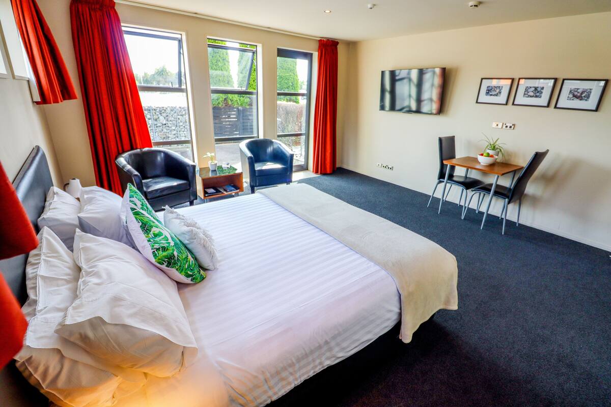 A hotel room with a large bed, a desk with chairs, a TV on the wall, and a window with red curtains.