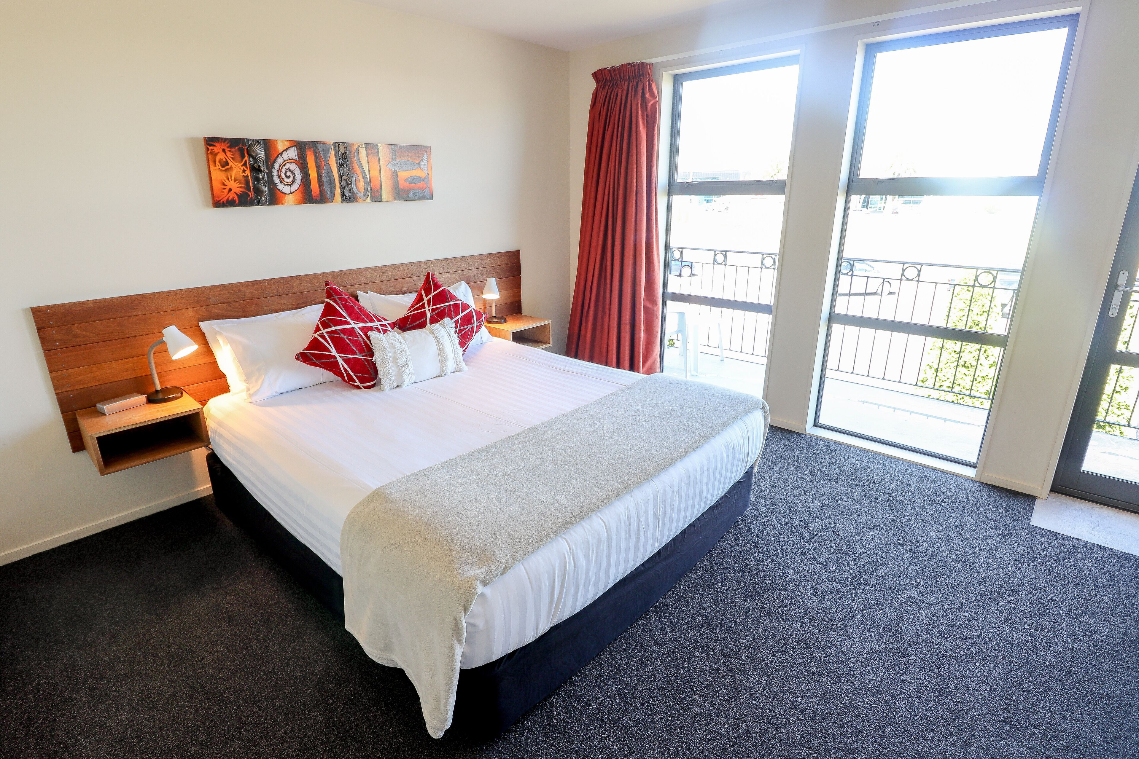 A hotel room with a large bed, a nightstand, a wall-mounted artwork, and a view through a window with a balcony.