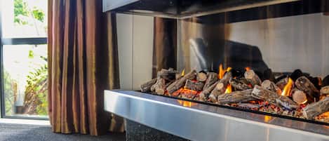 A modern fireplace with logs burning, a glass door, and a view of greenery outside.