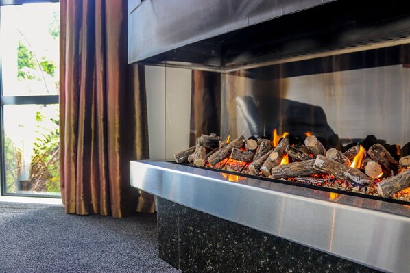 Una chimenea moderna con leña ardiendo, puerta de cristal y vista a la vegetación exterior.