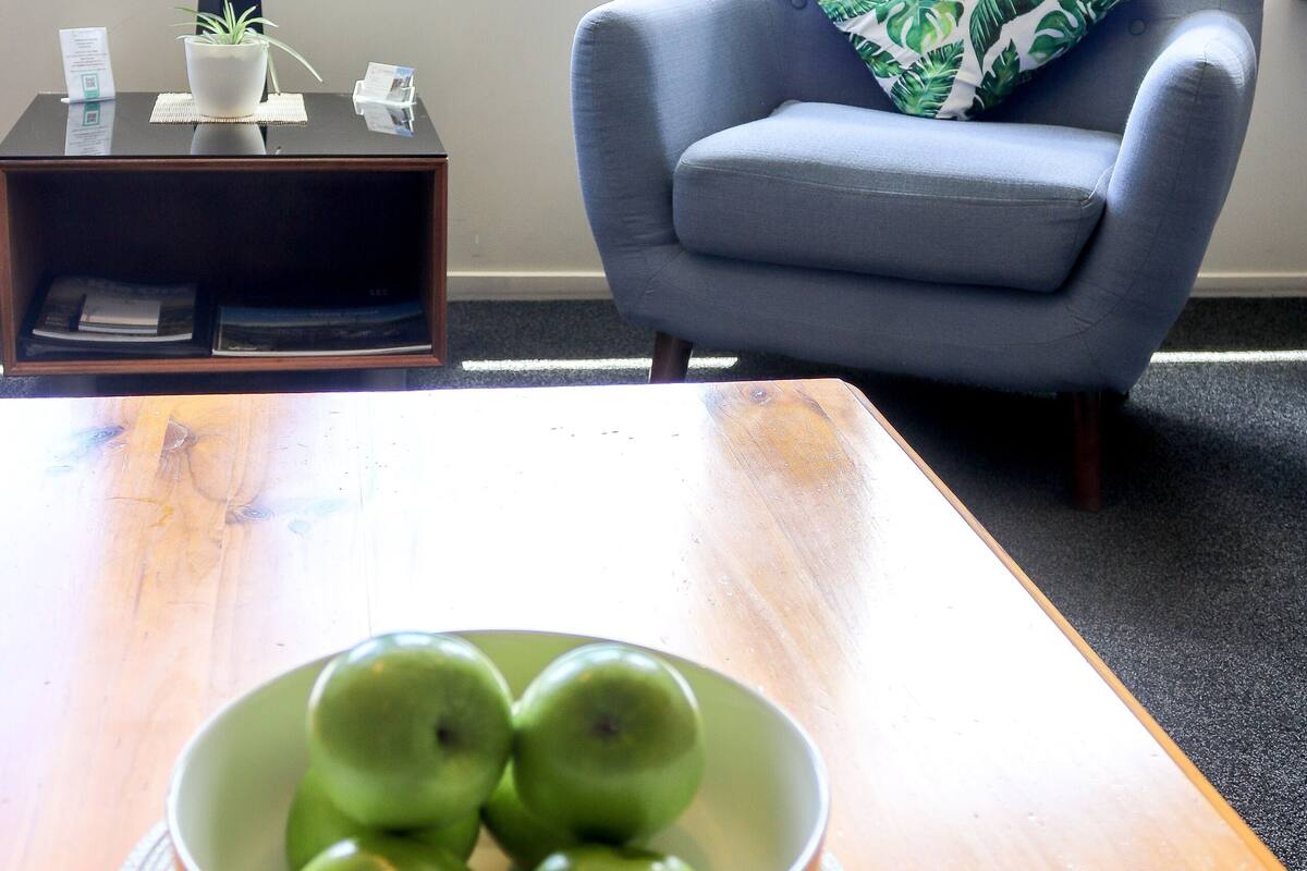 A room with a wooden table, a bowl of green apples, a blue armchair with a leaf-patterned pillow, a small side table with a lamp, and a window with a view of trees.