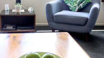 A room with a wooden table, a bowl of green apples, a blue armchair with a leaf-patterned pillow, a small side table with a lamp, and a window with a view of trees.