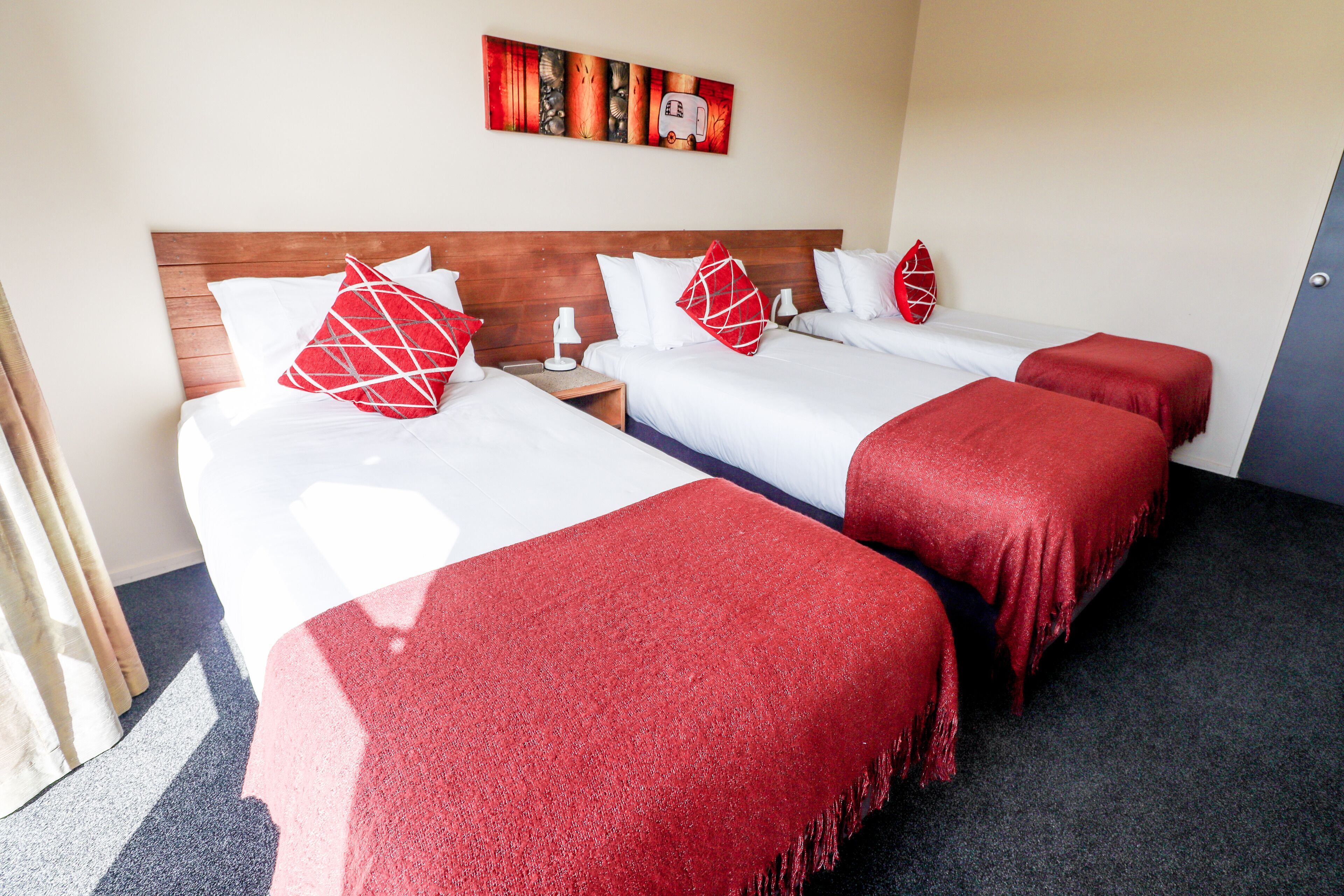 Habitación de hotel con dos camas, cada una con sábanas blancas y cojines rojos. Se observa un cabecero de madera, una mesita de noche con lámpara y un cuadro en la pared.