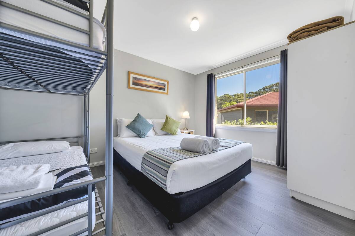 Habitación de hotel con literas, una cama individual, una ventana con vista a una casa y un cuadro en la pared.