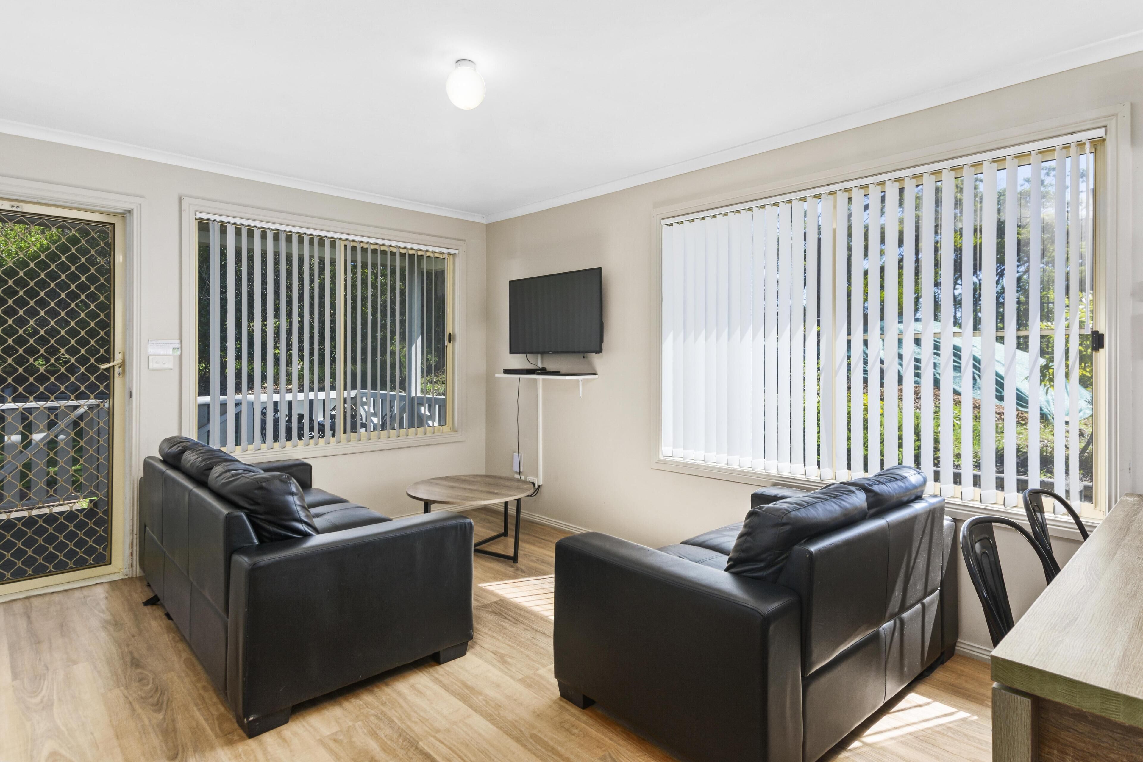 A modern living room with two black leather sofas, a flat-screen TV mounted on the wall, and large windows with blinds.