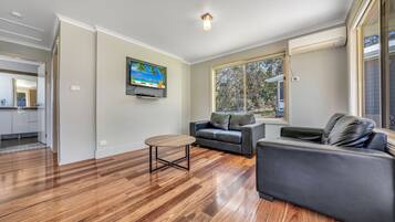 A spacious living room with hardwood flooring, a grey sofa, a coffee table, and a large window with a view of trees.