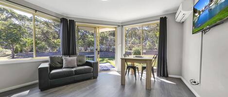 A modern living room with a sofa, dining table, and large windows offering a view of a garden.