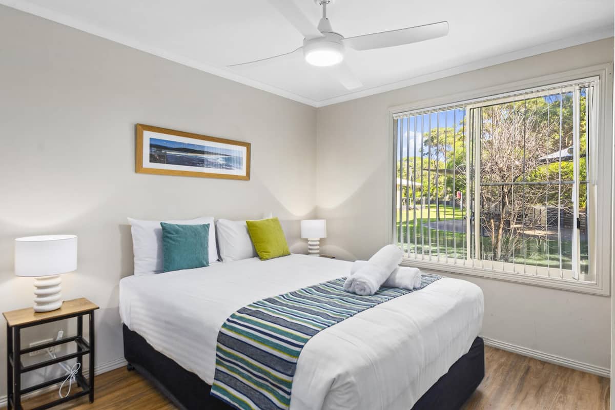 Habitación de hotel con una cama, ventana con vistas a zonas verdes, ventilador de techo y un cuadro en la pared.