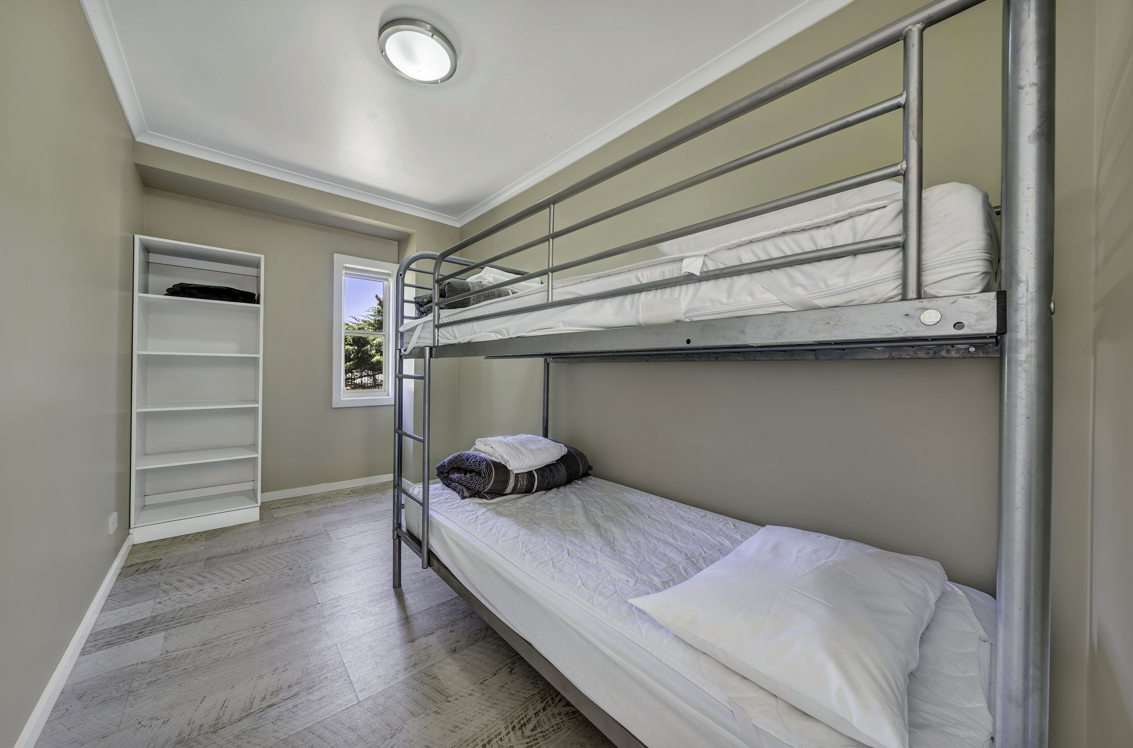 A bunk bed room with a metal ladder, a white mattress, and a white pillow. There is a white shelf unit against the wall and a door leading to another room.
