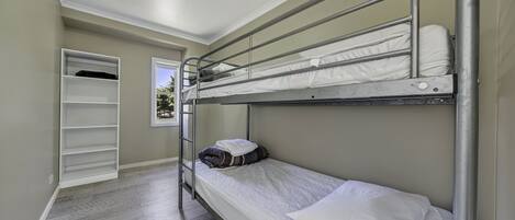A bunk bed room with a metal ladder, a white mattress, and a white pillow. There is a white shelf unit against the wall and a door leading to another room.